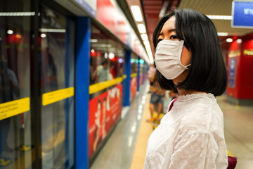 young adult Asian woman wearing a face mask on subway playform waitinf for train in Bangkok,...