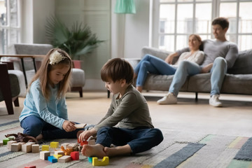 Joyful little baby girl playing toys on floor with small adorable brother while young couple parents resting on sofa in living room. Happy preschool siblings enjoying playtime at home at weekend.