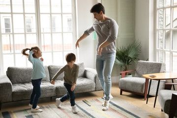 Loving young daddy having fun with cute kids siblings in living room, happy small brother and sister dancing to crazy disco music with energetic father at home, weekend family hobby activity concept.