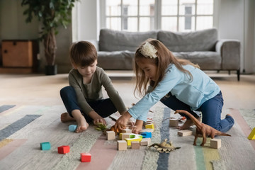 Cheerful little kids siblings sitting on warm floor, enjoying playing with cubes and toys in living room. Happy small brother and sister best friends having fun together without parents indoors.