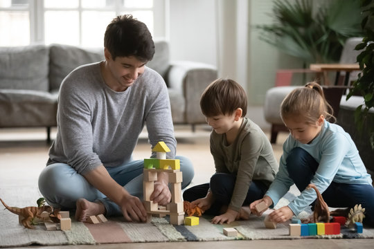 Caring young daddy sitting on floor, playing toys with lovely cute small daughter and son. Happy little children siblings having fun, enjoying playtime with affectionate loving father in living room.