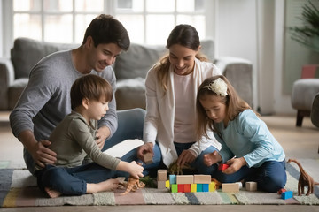 Happy couple parents playing with small preschool children on warm floor in living room. Joyful family of four enjoying spending weekend free leisure time together, constructing building with cubes.