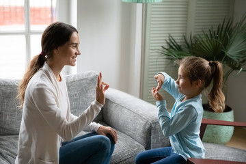 Smiling young mixed race woman teaching little cute girl sign language, showing symbols with fingers. Smiling hearing loss deaf disabled child communicating nonverbal with mommy in living room.