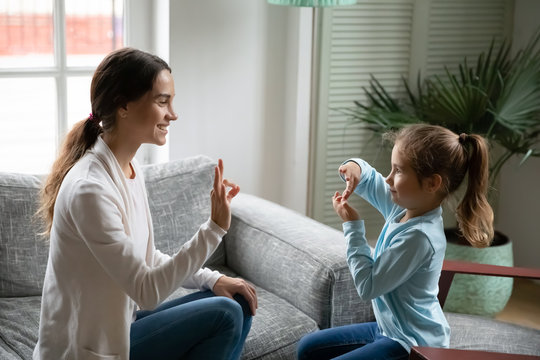 Smiling Young Mixed Race Woman Teaching Little Cute Girl Sign Language, Showing Symbols With Fingers. Smiling Hearing Loss Deaf Disabled Child Communicating Nonverbal With Mommy In Living Room.