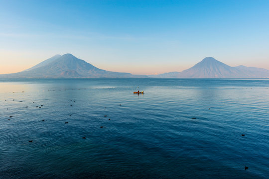 The volcanoes San Pedro, Toliman and Atitlan at sunrise by the Atitlan lake with a fisherman in a fishing boat between coots (Fulica atra), Panajachel, Guatemala.