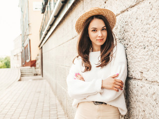 Portrait of beautiful cute model. Female dressed in warm hipster white sweater and hat. Trendy girl posing near wall in the street. Funny and positive woman hugging herself