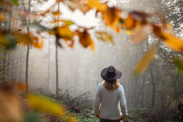 Fototapeta premium Moody atmosphere in autumn forest. Woman wearing knitted sweater and hat standing in fog at woodland
