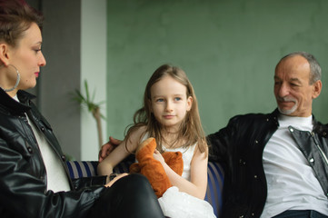 Family portrait of adult daughter, little granddaughter and senior grandfather in loft room with houseplants. Man and woman are wearing black leather jackets in punk style, generation concept