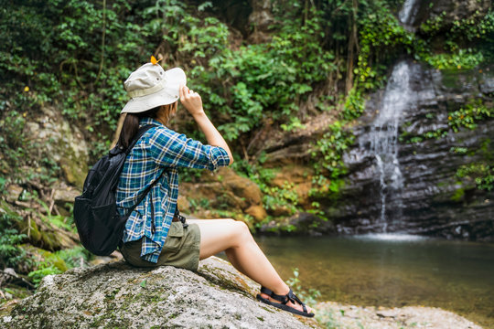 Asian girl traveler hiking traveling sitting on a rock with butterfly on hat exploring jungle nature forest waterfall, wearing travel gear, summer hot healthy outdoor peaceful freedom lifestyle