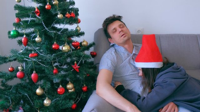Couple man and woman sleeping together on couch near Christmas tree at home. New Year night on winter holidays. Morning hangover after party. Family life. Resting hugging each other.