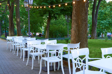 open air street cafe outdoor - white chairs and tables, decorated with illumination, the city Park...