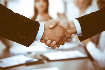 Business people shaking hands at meeting while theirs colleagues clapping and applauding. Group of unknown businessmen and women in modern white office. Success teamwork, partnership and handshake