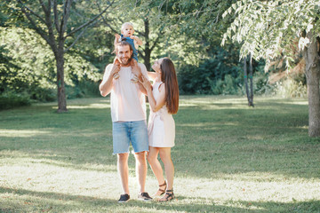 Naklejka na ściany i meble Happy smiling mother and father with baby boy in park outdoors. Family Caucasian mom and dad with son walking hugging in park on summer day. Happy family authentic lifestyle concept.
