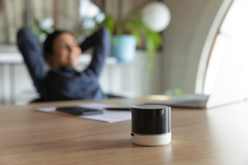 Close up focus on portable stylish little digital assistant on table, with relaxed businesswoman on background. Happy worker using small voice speaker or enjoying pleasant music from column in office.