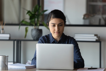 Focused young indian ethnicity businesswoman sitting at table with computer, working alone on...