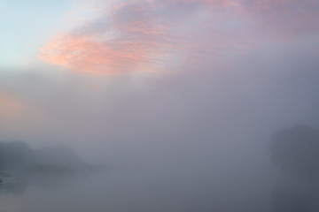 Tranquil morning landscape at sunrise. Heavy fog over the river. Dawn illuminates and makes the clouds colorful.