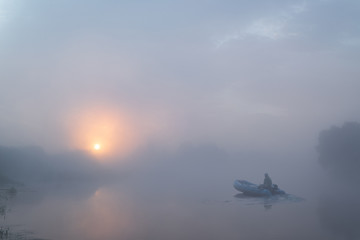 Fototapeta na wymiar Tranquil morning landscape panorama at sunrise. A fisherman on a rubber boat on the river in heavy fog. Dawn illuminates and makes the clouds colorful.