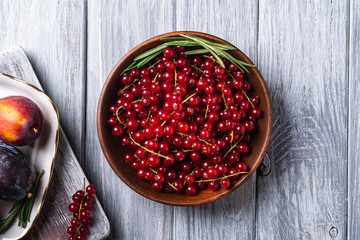 Fresh red currant berries in wooden bowl near to sweet plum fruits in plate with rosemary leaves on old cutting board, grey wood background, top view