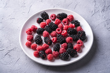 Tasty fresh ripe raspberry and blackberry berries in plate, healthy food fruit on stone concrete background, angle view
