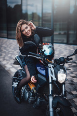 Fototapeta na wymiar Young cute female driver and dark modern bike. Urban powerfyl vehicle. Motosport hobby.