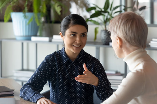 Friendly millennial indian businesswoman talking to blonde female colleague, sitting at table in office. Two young multiracial employees discussing working issues or enjoying informal conversation.