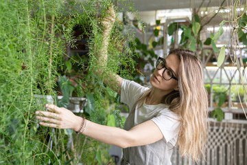 Joyful young woman gardener in lien dress touching lush asparagus fern houseplant. Greenery at home. Love of plants. Indoor cozy garden. 