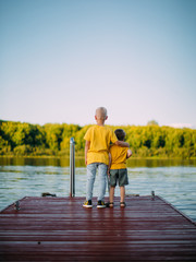 Cool kids brothers hug each other and look at the river staying on dock. Back view