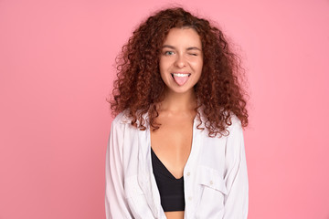 Pretty cheerful redhead girl with curly hair showing tongue at camera over pink background.