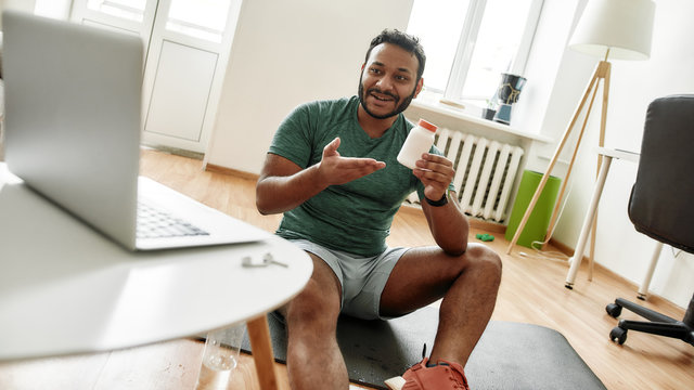 Nutrition and diet. Male fitness instructor smiling, advertising sport nutrition while streaming, broadcasting video lesson on training at home using laptop. Online personal trainer