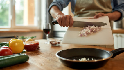 Close up of man in apron putting onion in the pan for frying while cooking dinner. Healthy nutrition, cooking at home concept