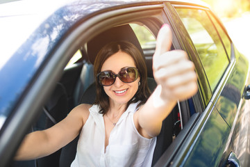 A smart looking middle-aged adult woman wearing sunglasses put her hand out the car window and raised her thumbs up. smiling woman drives Car