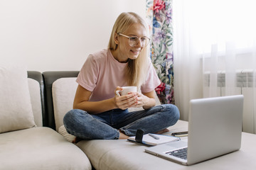 Happy young woman sit on couch in living room browsing surfing wireless internet on laptop, smiling millennial girl rest on sofa texting messaging on computer at home, shopping online via web
