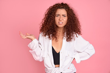 Young displeased redhead girl gesturing looking at camera. Confused girl arguing with a boyfriend. Pink background.