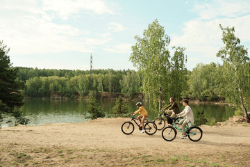 Side view of young family sitting on bicycles and moving along riverbank