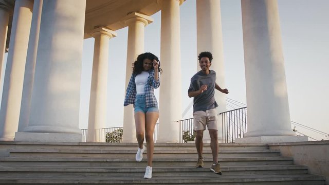 Dark-skinned pair laughing and dancing while go downstairs from pillared observation deck in park