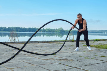 athletic man in the morning, against the background of the lake doing exercises with gymnastic ropes