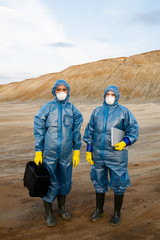 Two young female researchers in protective workwear standing on polluted soil