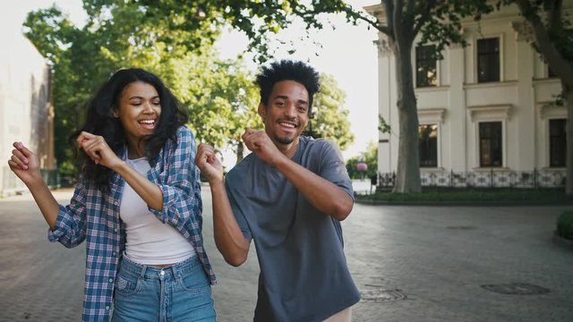 Ethnic black male and female smiling, dancing synchronously while walking by city square