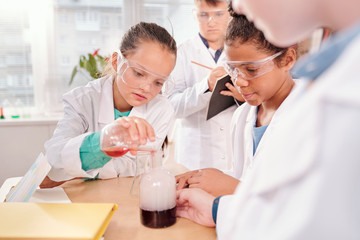 Cute schoolgirl mixing two fluids in bottle while one of classmates writing