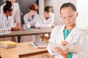 Fototapeta premium Serious pupil of secondary school in whitecoat and eyeglasses making notes