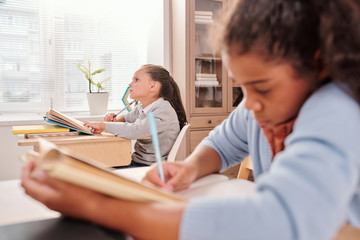 Pensive schoolgirl with open book thinking of answer to question on blackboard