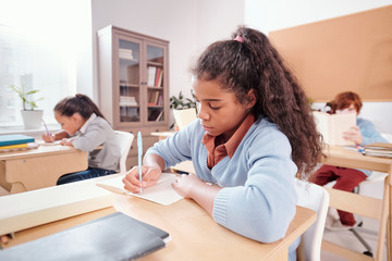 Serious mixed-race schoolgirl making notes or wtiting essay in copybook
