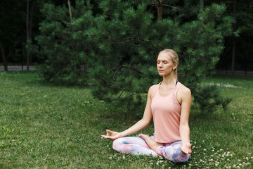 Beautiful young woman seating in Yoga pose of lotus in the park and meditating on green grass