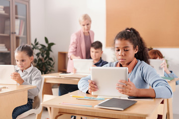 Group of youthful contemporary schoolchildren with touchpads sitting by desks