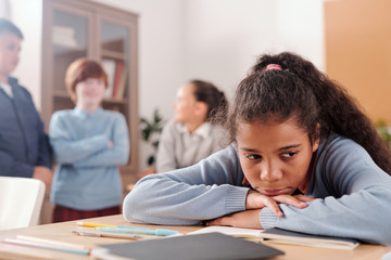 Upset or offended schoolgirl sitting by desk against classmates talking