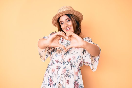 Young beautiful caucasian woman wearing summer dress and hat smiling in love doing heart symbol shape with hands. romantic concept.