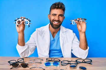 Young hispanic man with optometry glasses sitting at the table winking looking at the camera with...