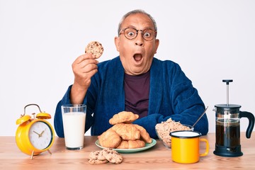 Senior handsome man with gray hair sitting on the table eating breakfast in the morning scared and amazed with open mouth for surprise, disbelief face