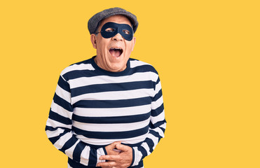 Senior handsome man wearing burglar mask and t-shirt smiling and laughing hard out loud because funny crazy joke with hands on body.