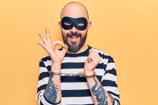 Young handsome man wearing burglar mask and handcuffs doing ok sign with fingers, smiling friendly gesturing excellent symbol